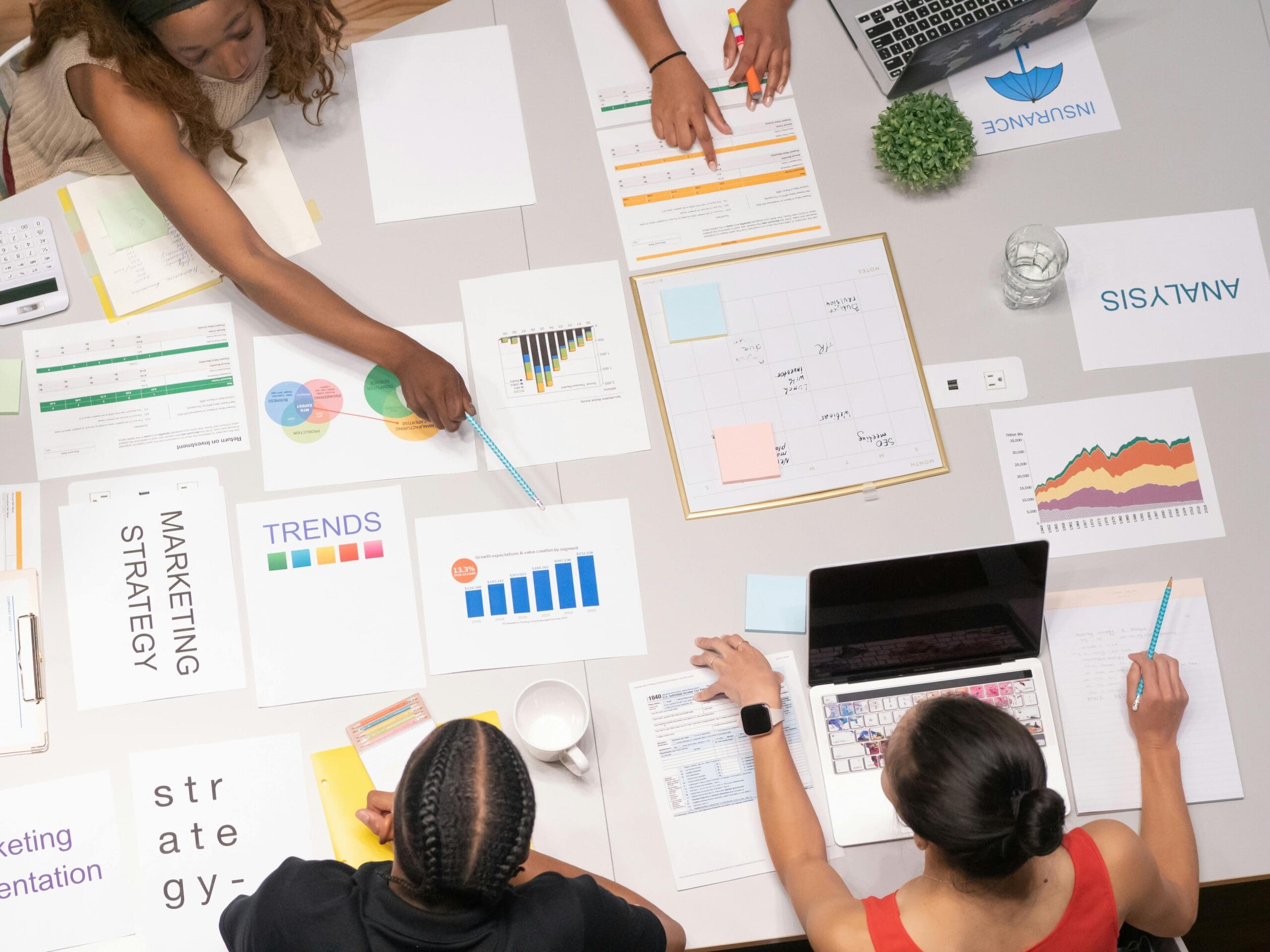 Team of three women and one man collaborating around a table covered with marketing strategy documents, charts, and analytics reports, with laptops and colorful data visualizations spread across the workspace.